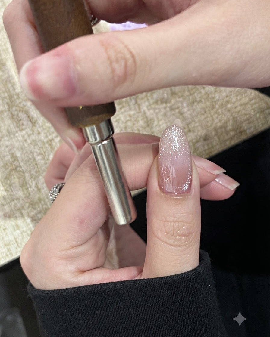 Una mano che tiene un magnete cilindrico in orizzontale sulla punta di ununghia dipinta con gel cat-eye scintillante, non ancora polimerizzato.