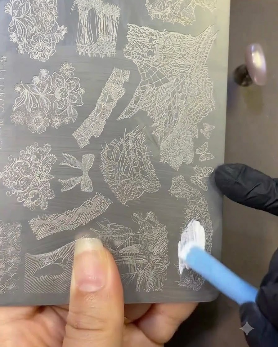 Un pennello che applica lo smalto bianco per stamping su una piastra metallica incisa con motivi in pizzo.