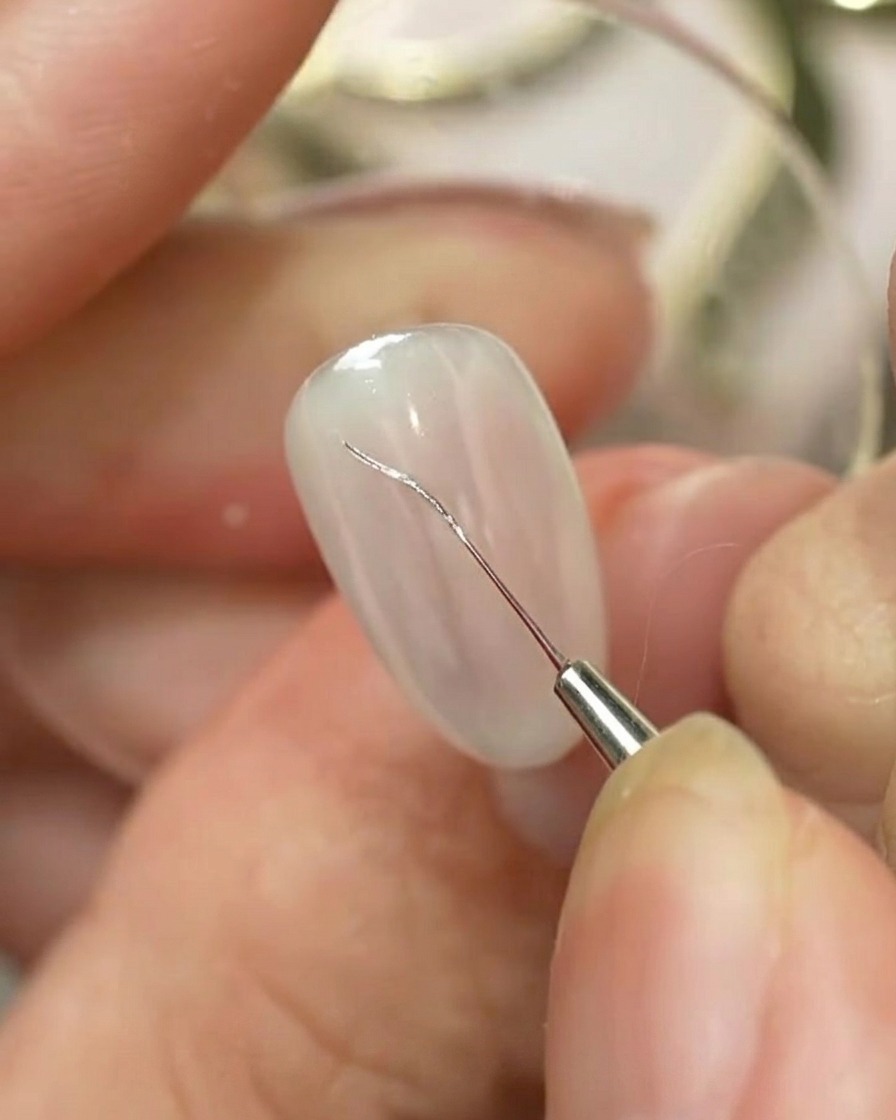 Un pennello liner fine per nail art che disegna un contorno argentato di un fiocco su unghie curate di colore chiaro.