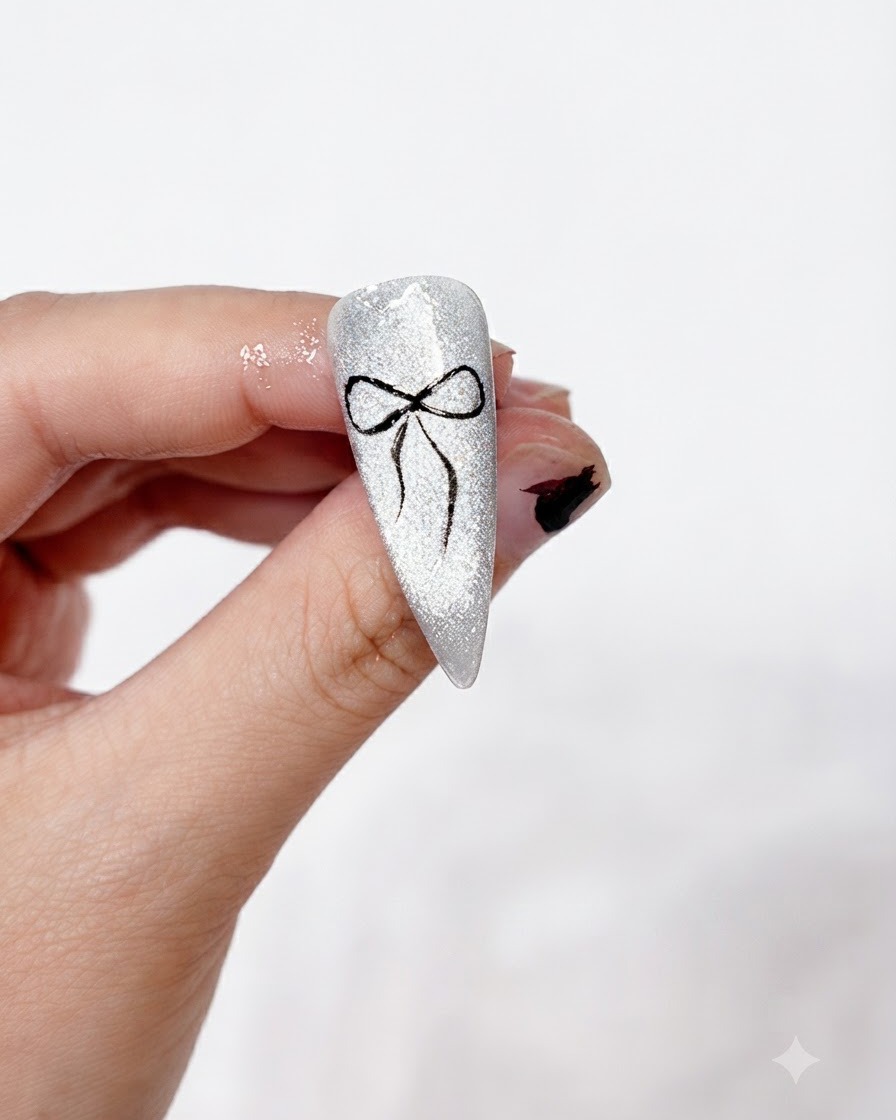 Un design di nail art con fiocco nero completato, dipinto in modo pulito e impostato su uno sfondo cat-eye argento scintillante, tenuto tra le dita.