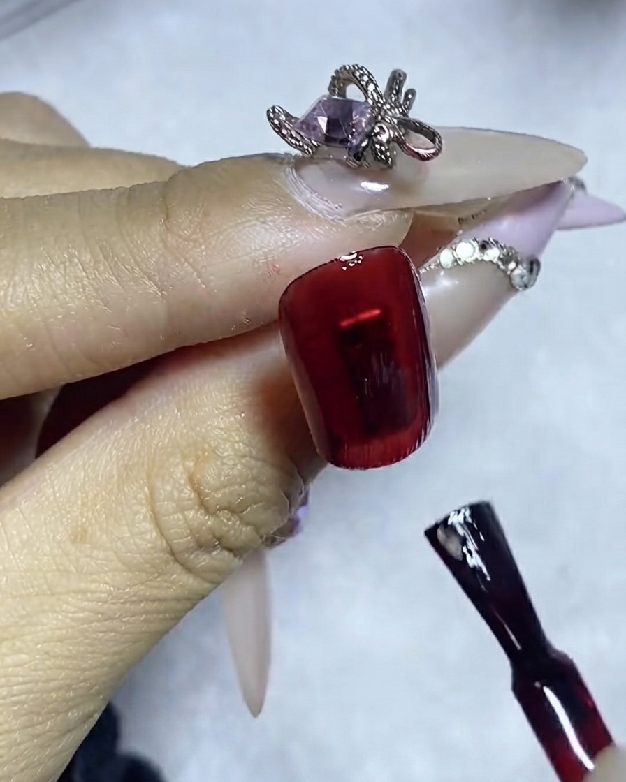 Un pennello liner che applica una linea precisa di colla per gemme su una superficie di unghia smaltata di rosso scuro.