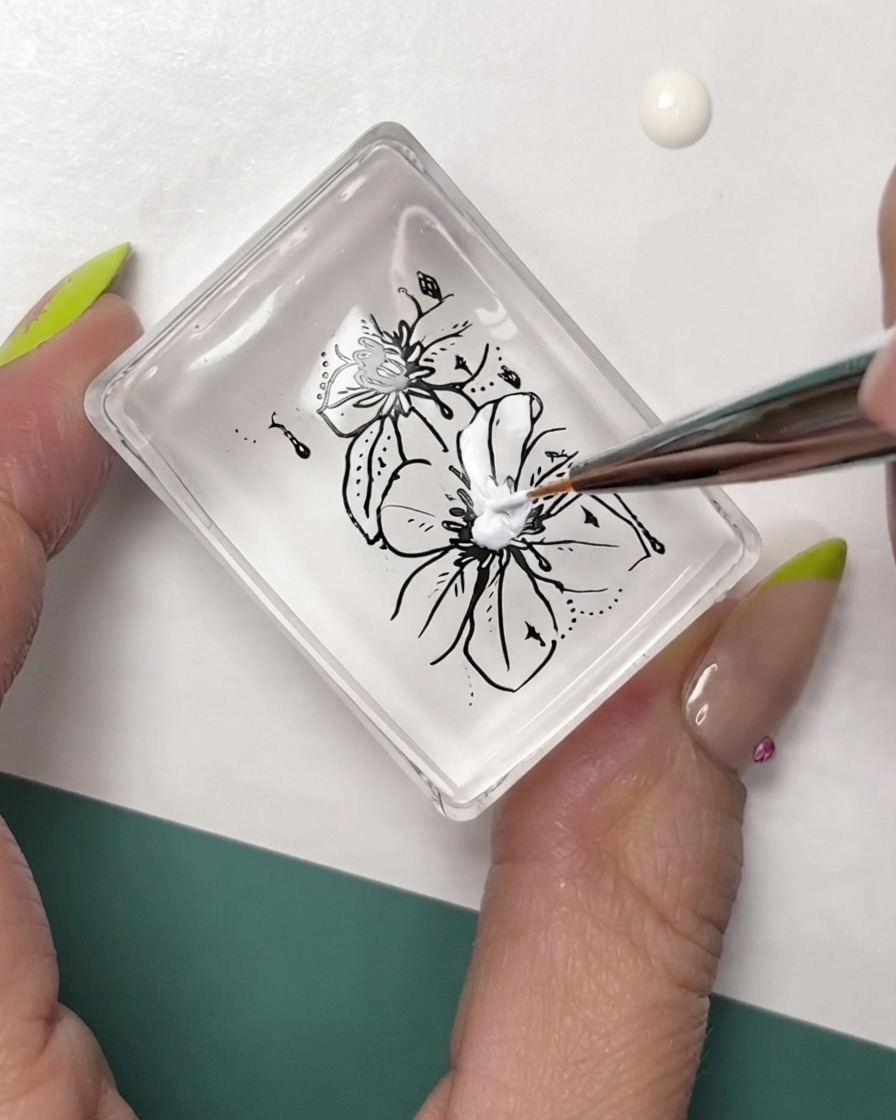 Un pennello sottile per nail art che aggiunge dettagli bianchi su una decorazione floreale con contorno nero applicata su un cuscinetto per timbro trasparente.