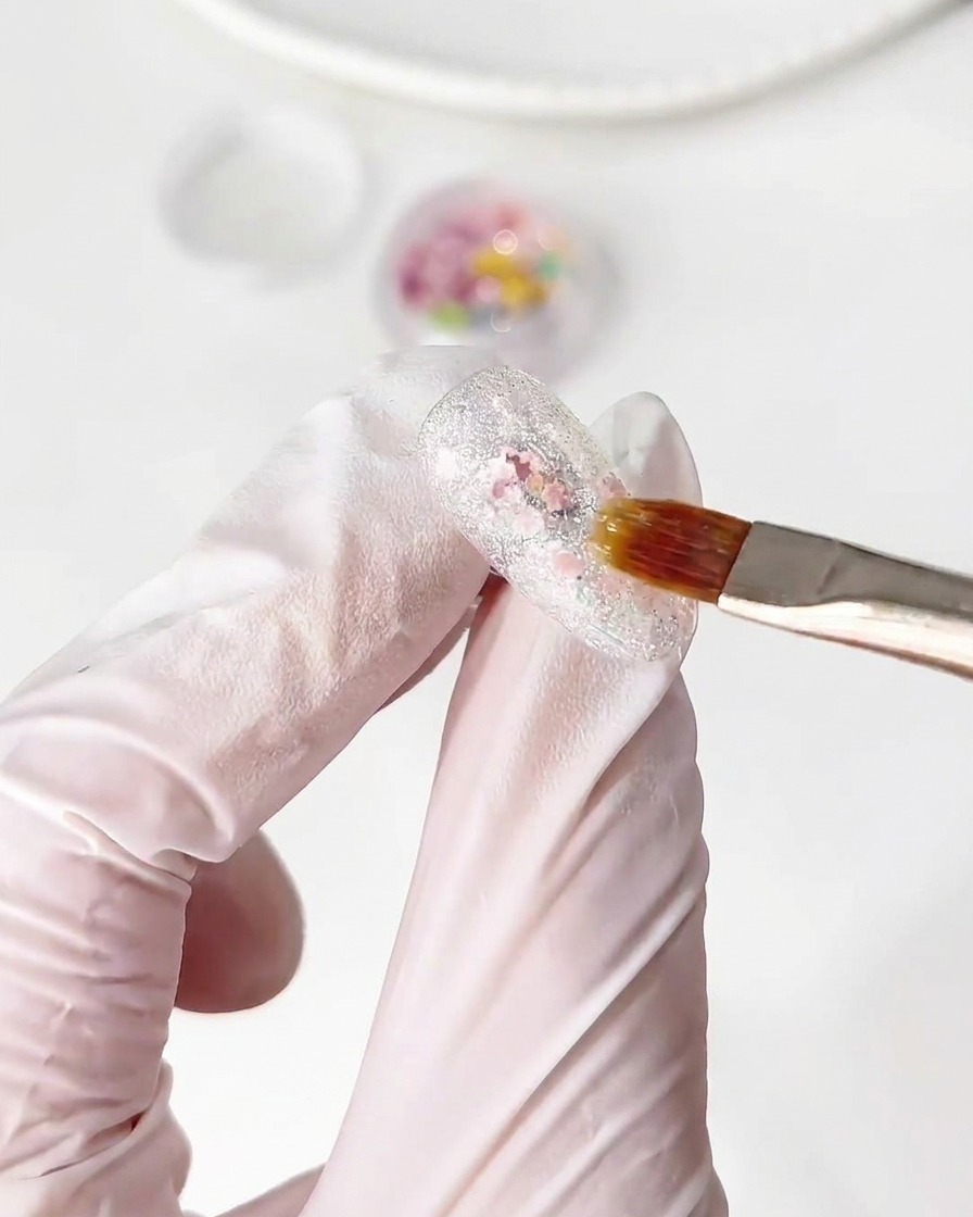 Un pennello posiziona piccoli pezzi di glitter floreali colorati su uno strato di base gel umido su ununghia.