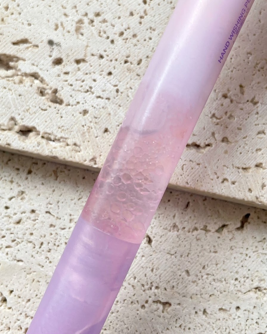 Una penna per olio per cuticole con essenza di perla dal gradiente rosa tenuta contro uno sfondo strutturato.