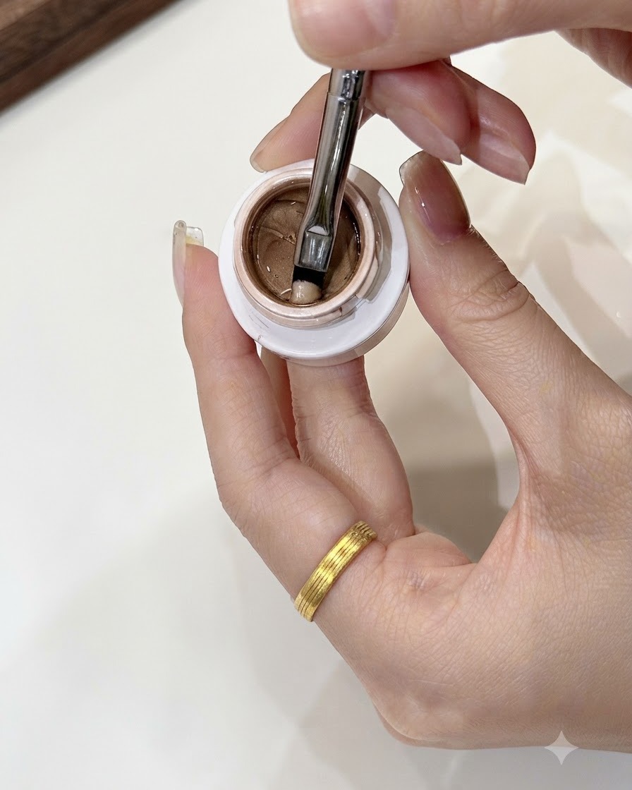Un pennello da nail art che preleva una piccola porzione della misura di un fagiolo mung di gel oro champagne da un barattolo aperto.