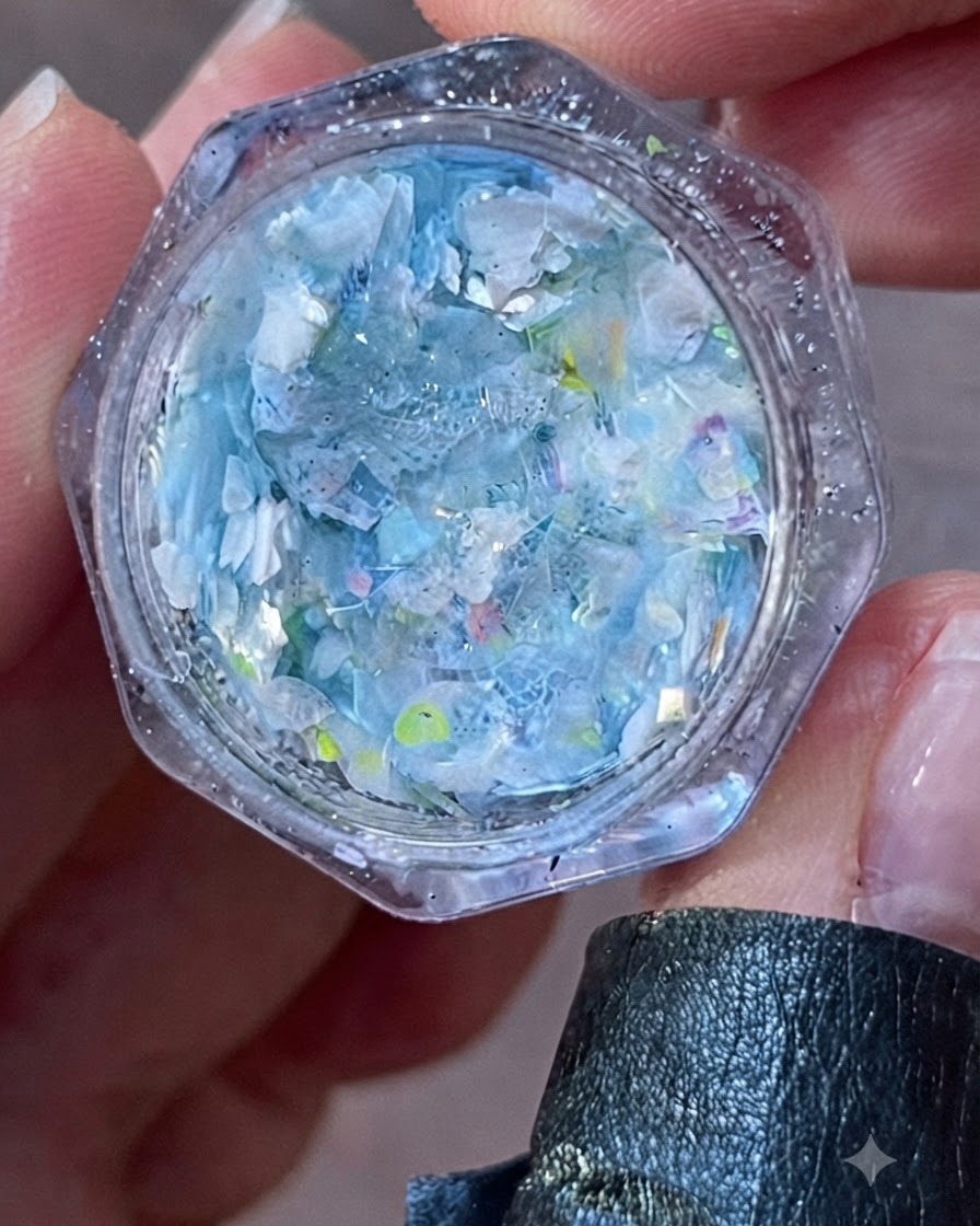 Primo piano di un barattolo aperto pieno di scaglie e glitter iridescenti blu e bianchi.