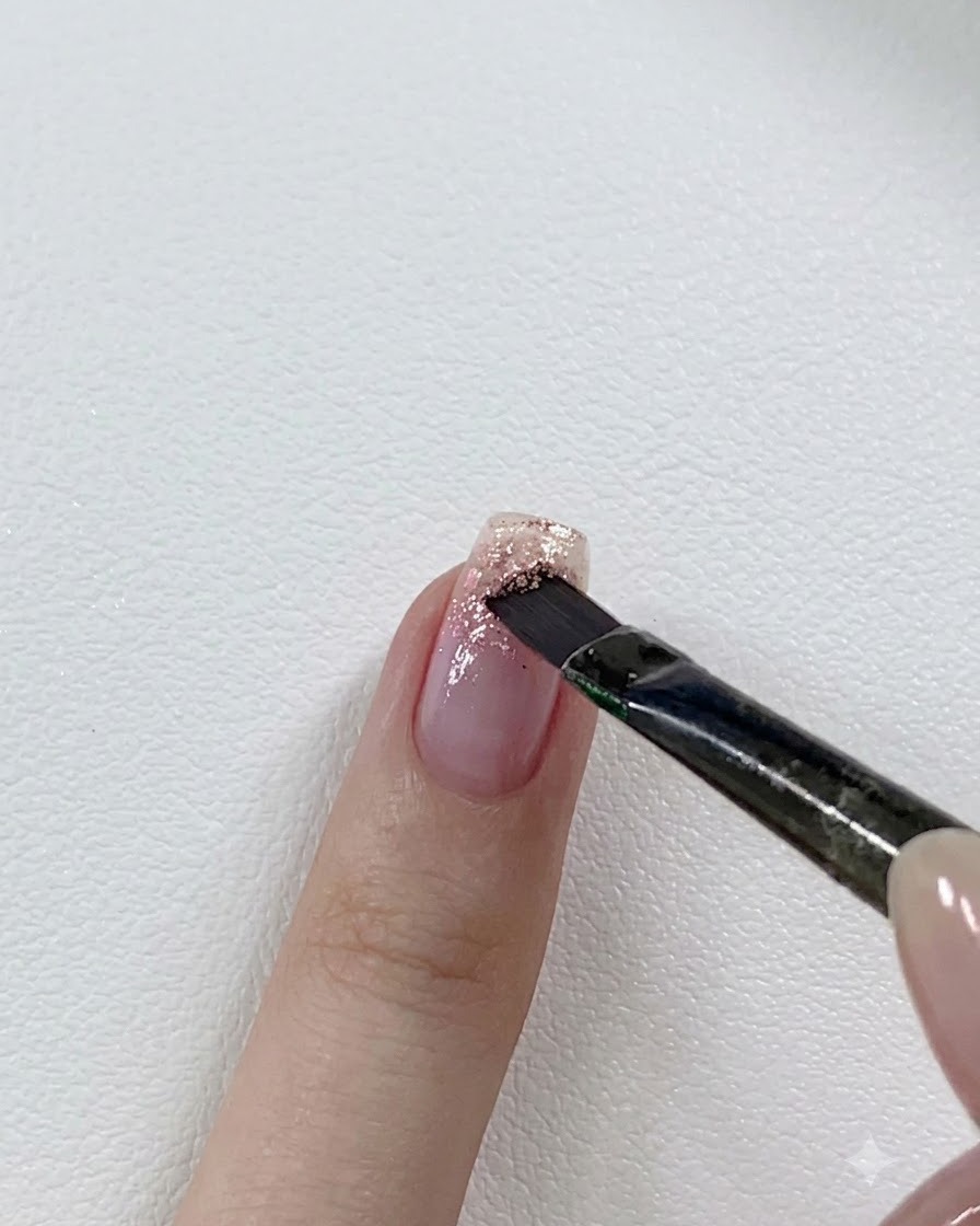 Un pennello per unghie stratifica un gel glitter rosa più scuro sopra la punta di ununghia parzialmente glitterata.