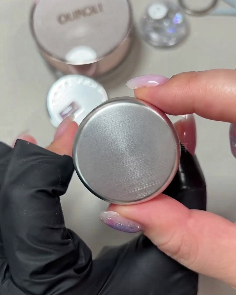 Una mano che mostra un piccolo barattolo rotondo di smalto gel cat-eye bianco porcellana con riflessi su sfondo neutro.