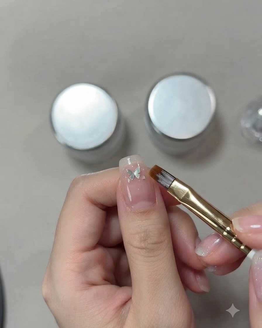 Un primo piano dettagliato della mano di una nail artist che utilizza un pennello sottile per posizionare una piccola paillettes a farfalla argentata su ununghia cat-eye rosa.