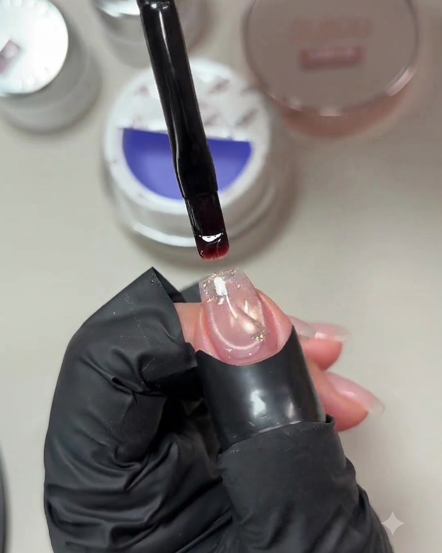 Un pennello che trattiene una goccia di gel costruttore viola-blu tenue sopra ununghia preparata con una paillettes incorporata.