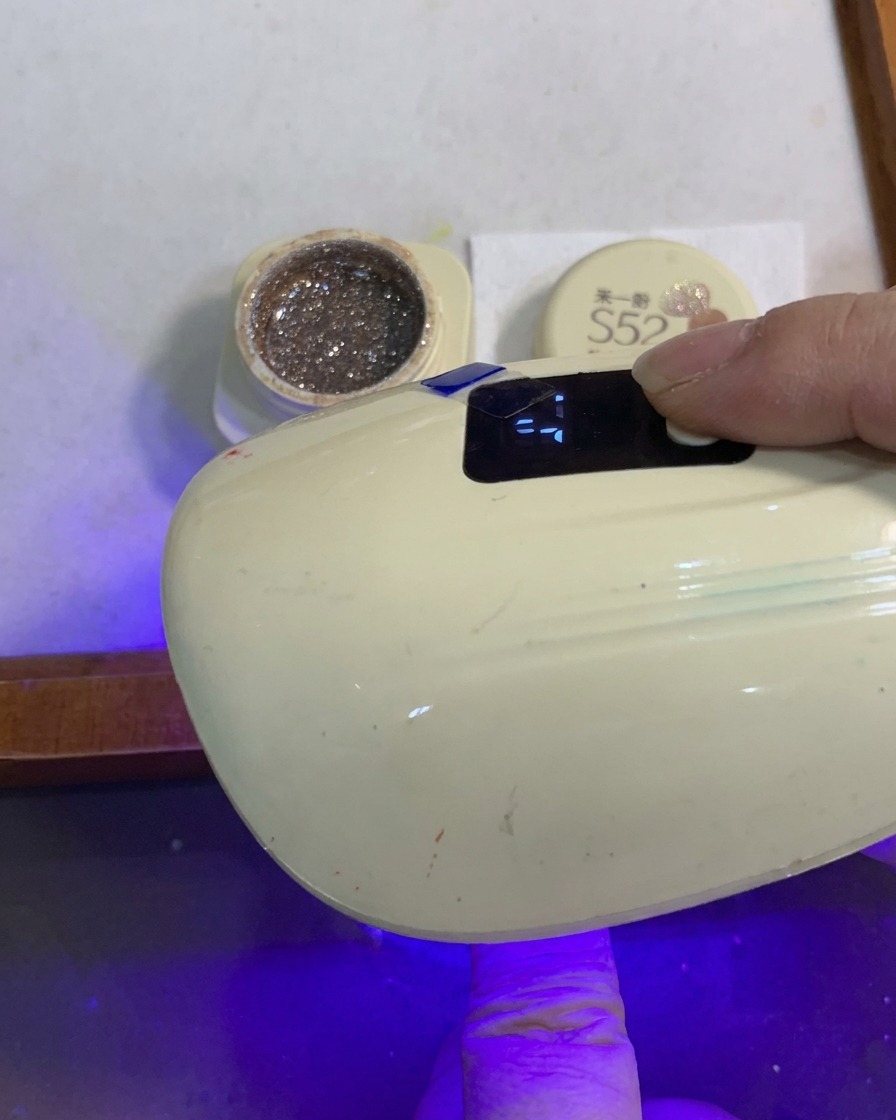 Un dito con ununghia sfumata di glitter sotto una piccola lampada UV/LED.