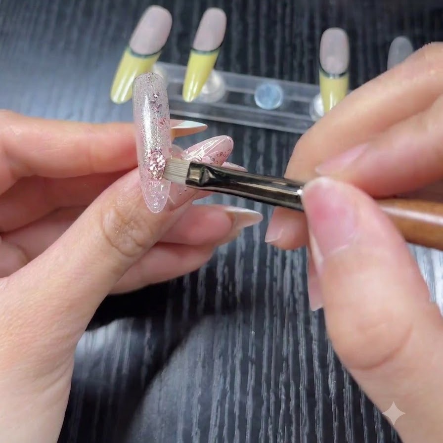 Un primo piano di un pennello che applica scaglie di glitter olografici rosa e argento su una base semitrasparente su una tip da pratica.