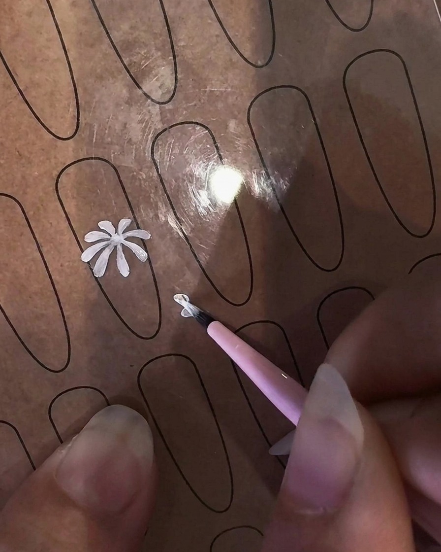 Primo piano di una mano che impugna un pennello liner fine per dipingere un petalo di margherita bianco su un foglio trasparente per pratica unghie.