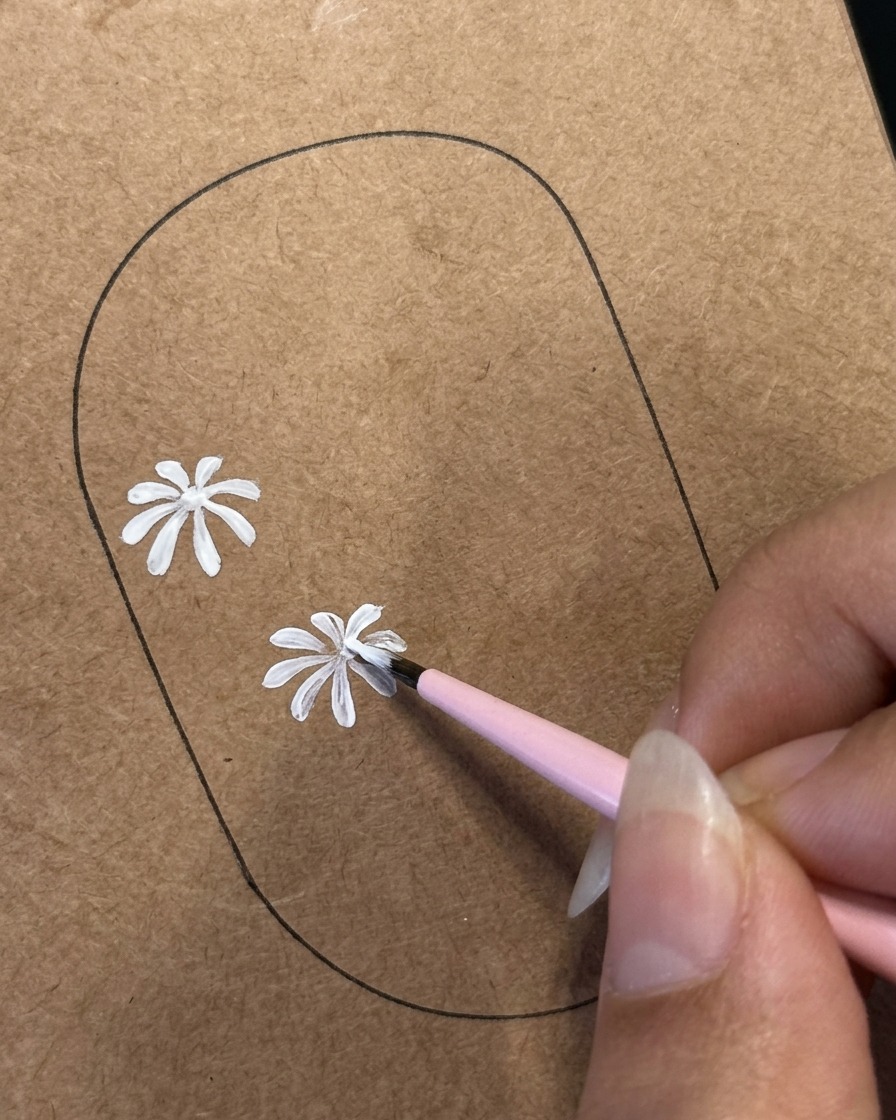 Un pennello liner fine che dettaglia i petali bianchi su un secondo design di margherita dipinto su un modello marrone di pratica per unghie.