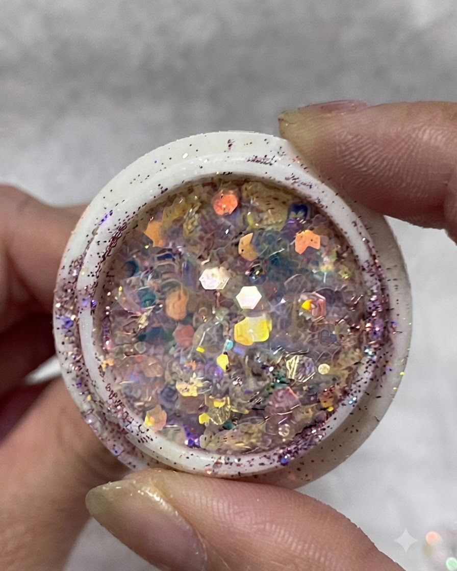 Un vasetto pieno di fiocchi glitter iridescenti multicolore, paillettes e piccole stelle tenuto tra le dita.