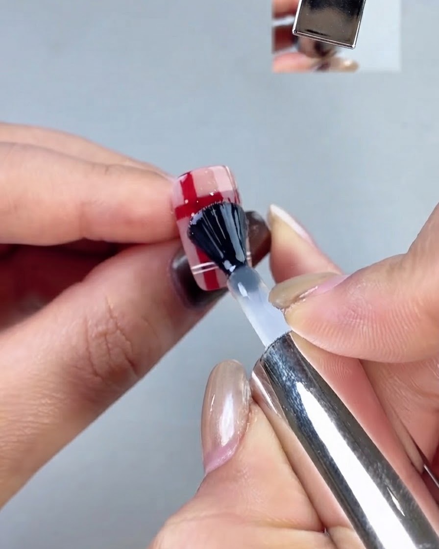 Un pennello per unghie nero che applica uno strato di top coat trasparente su un design scozzese.