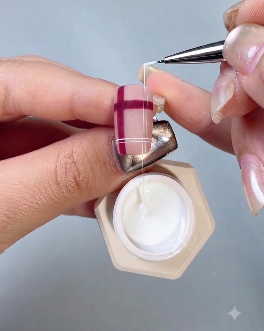 Un pennello fine da nail art che disegna sottili linee bianche con gel seta sopra un motivo scozzese rosso su ununghia.