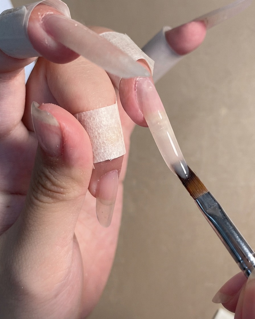 Un pennello da unghie che applica un gel nude semitrasparente su unestensione di unghia stiletto lunga.