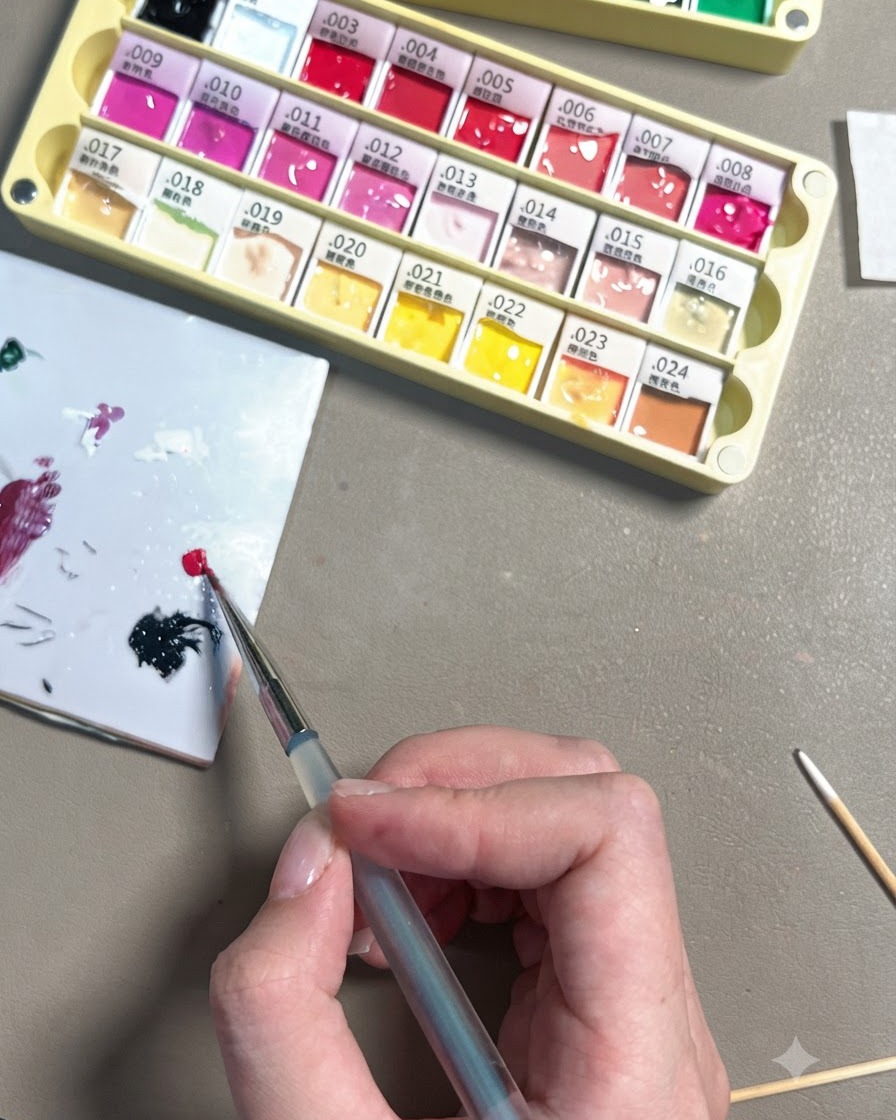 Una mano che usa un pennello fine da dettaglio per depositare una piccola goccia di gel rosso rosa brillante su una tavolozza di ceramica bianca.