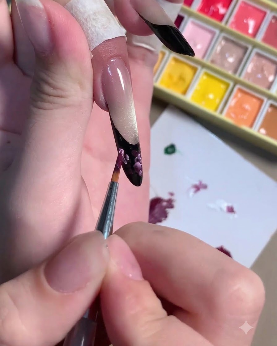 Un pennello fine che applica tratti di luce rosa chiaro sopra la nail art floreale prugna scuro.