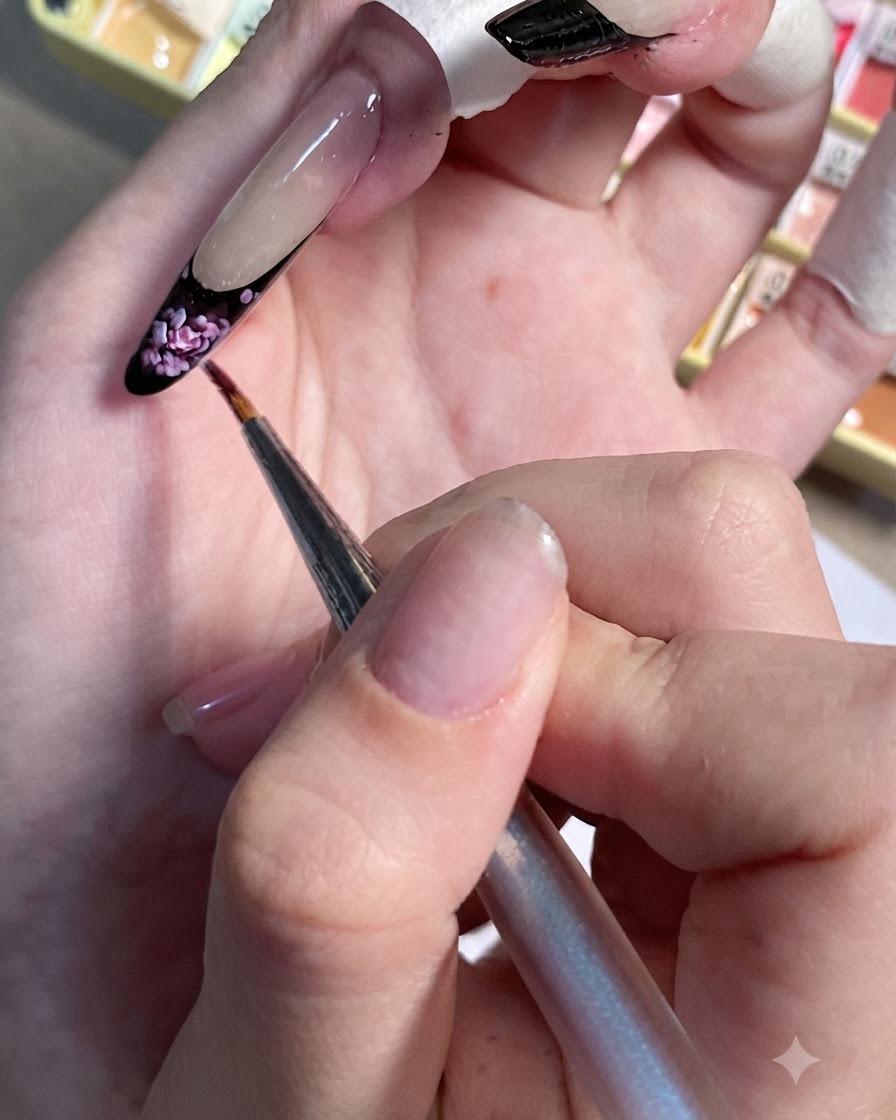 Un pennello di dettaglio che applica piccoli punti di rosa chiaro sulla French tip nera di ununghia dipinta.