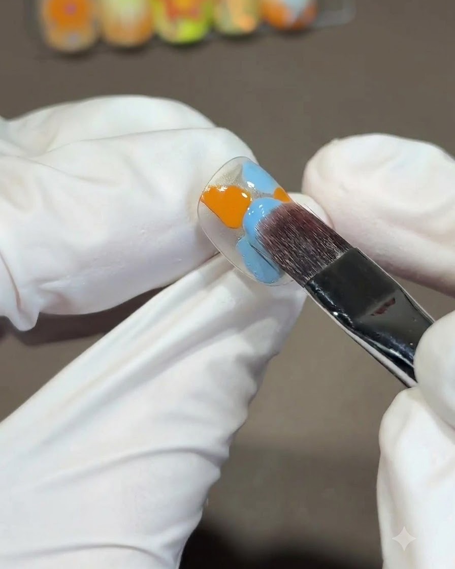 Un grande pennello quadrato che sfuma smalti gel arancioni e blu su una tip per unghie usando un movimento continuo a S verso il basso.