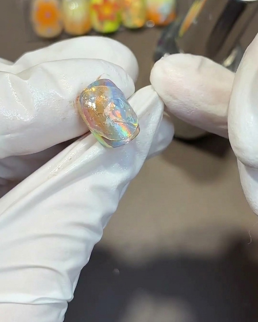 Lutilizzo di uno strumento di metallo per imprimere texture fluide e ondulate nel gel trasparente denso sopra una tip per unghie iridescente.