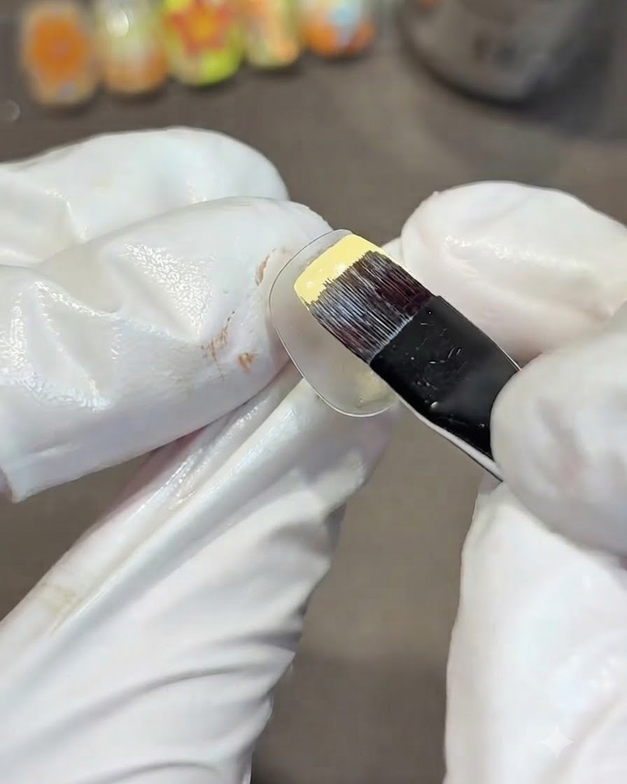 Una mano guantata che utilizza un pennello piatto per gel per dipingere ununghia pratica trasparente con smalto gel giallo solido.