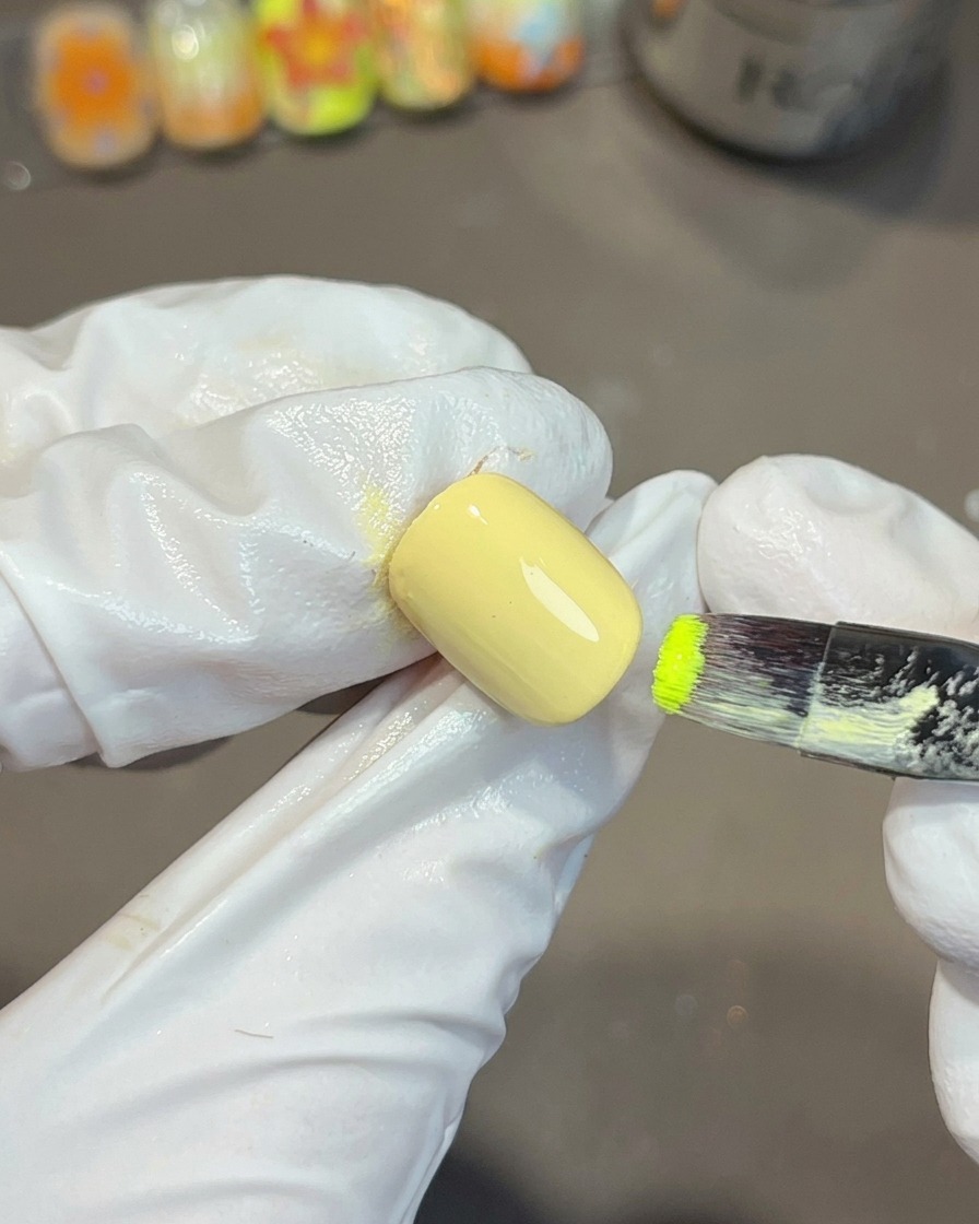 Una mano guantata che utilizza un pennello piatto per unghie per applicare smalto gel verde vibrante sullangolo di ununghia dipinta di giallo.