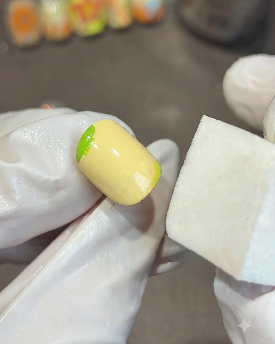 Una mano guantata che tiene un blocco di spugna bianca accanto a ununghia gialla, sfumando una sfumatura di gel verde sullangolo.