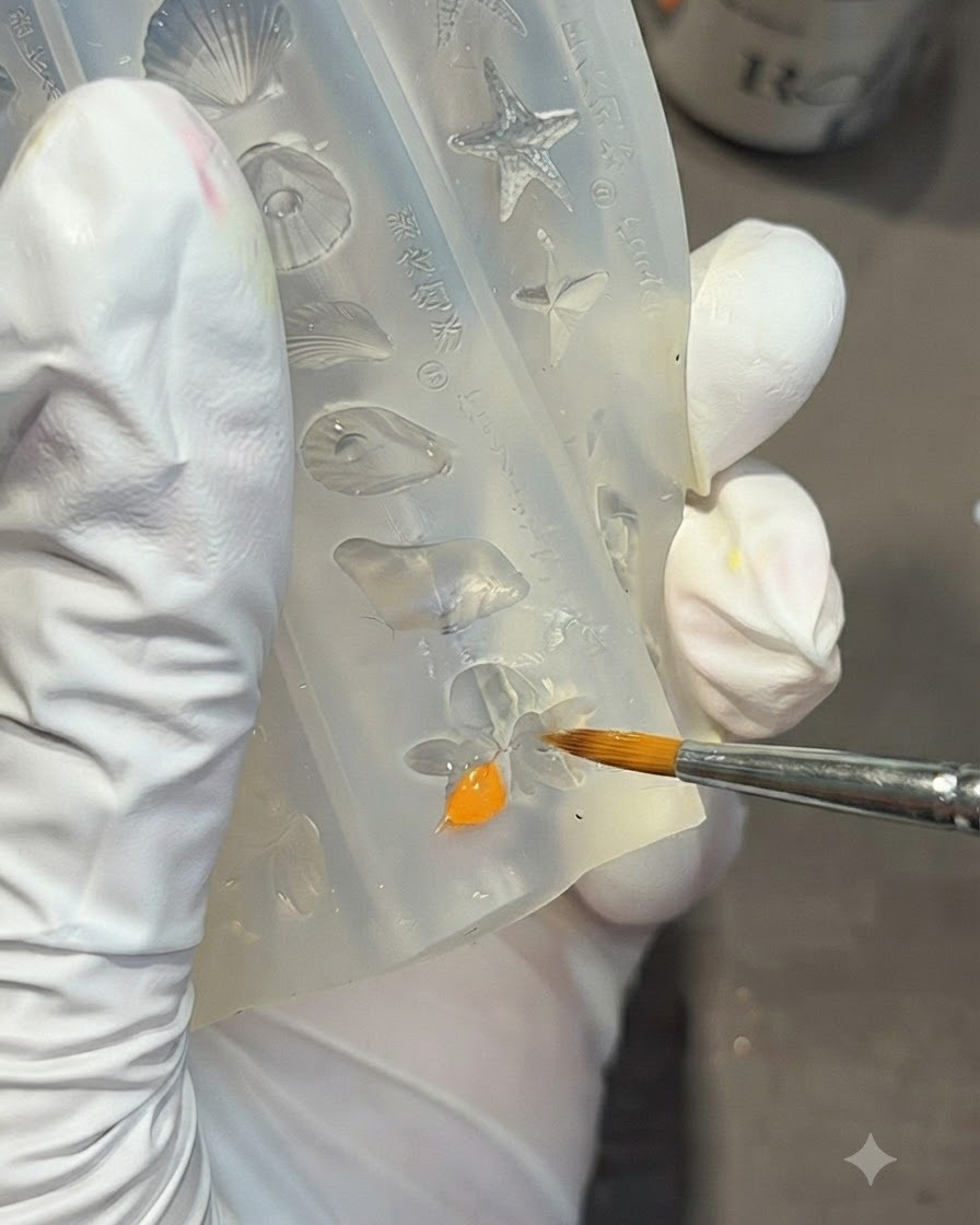 Una mano guantata che utilizza un pennello di dettaglio per riempire la forma di un fiore di giglio allinterno di uno stampo in silicone trasparente con gel arancione brillante.