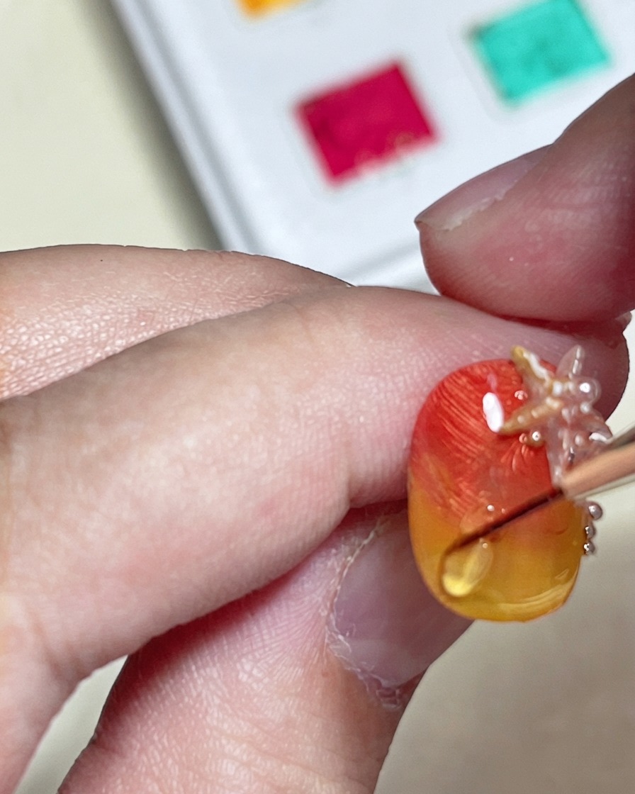 Un pennello che disegna una linea di increspatura dellacqua lucida e in rilievo con un hard top coat su ununghia con gradiente arancione e giallo materico.