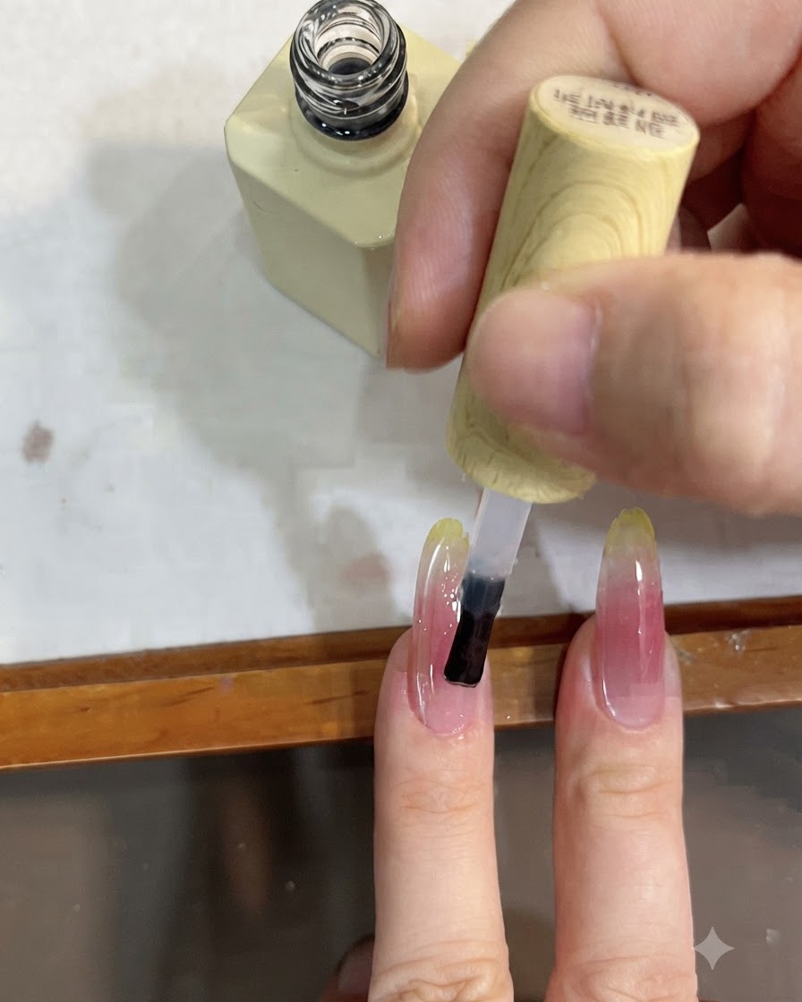 Un primo piano di un pennello che applica un top coat trasparente su ununghia lunga con sfumatura rosa e gialla.