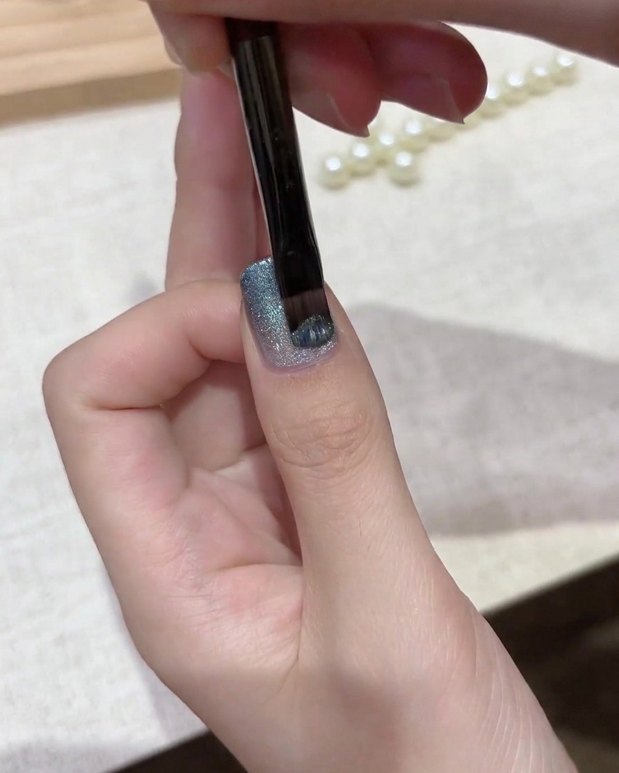 Un pennello per gel applica una mano di lucido magnetico cat eye argento-blu brillante su una base sfumata.