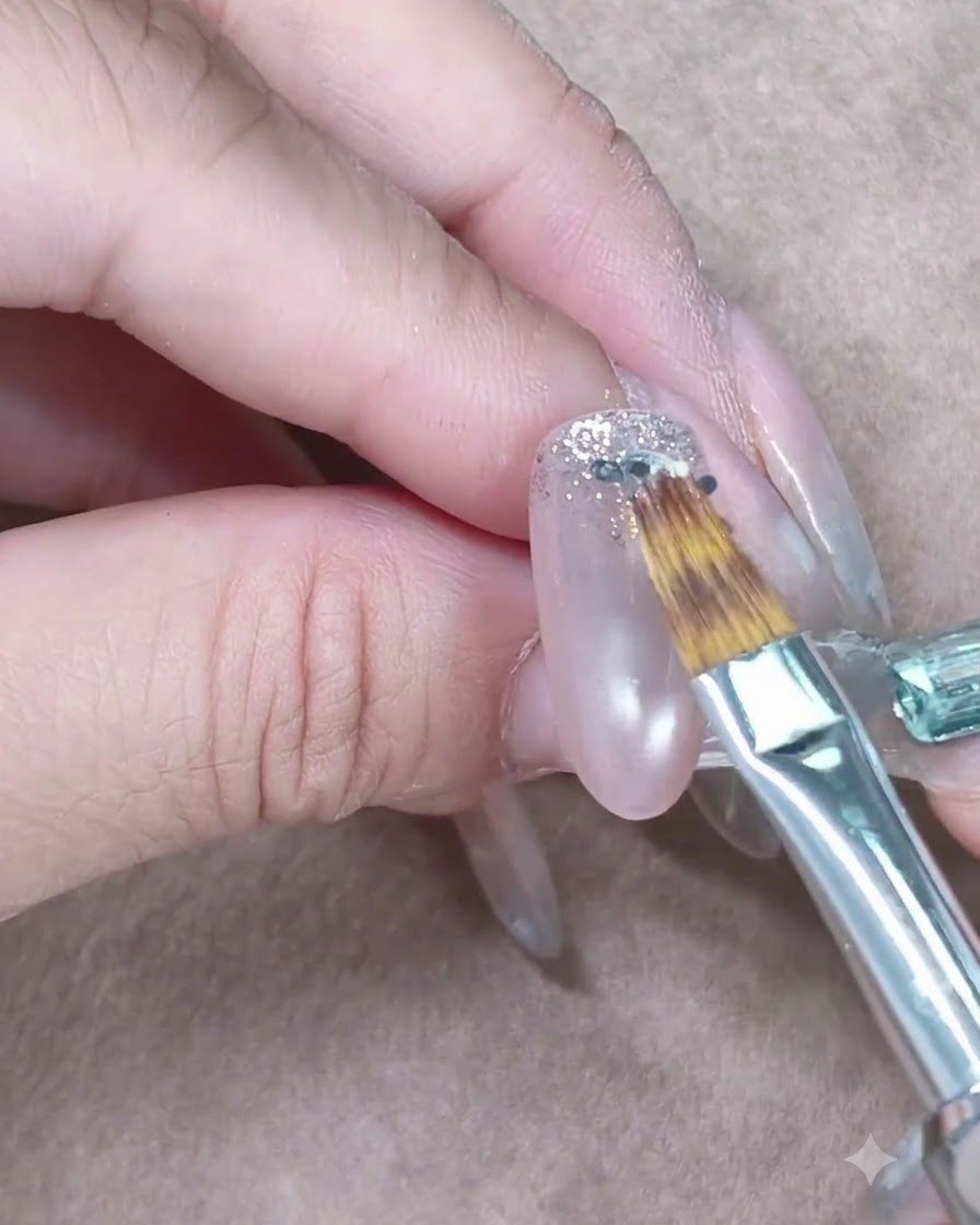 Un piccolo pennello da unghie che allunga e sfuma delicatamente i glitter argentati verso il basso su ununghia in gel trasparente.