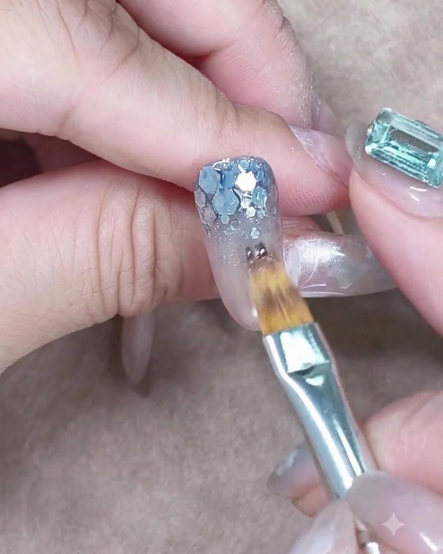 Un pennello piatto che tampona il gel blu trasparente sulle paillettes argentate grosse alla base dellunghia.