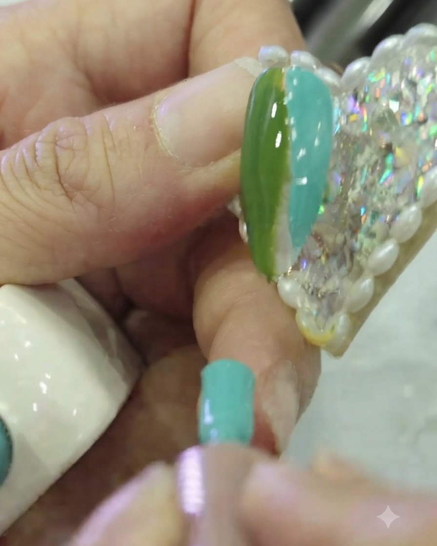 Primo piano di un pennello piatto per nail art che tampona il confine tra lo smalto gel verde scuro e teal su ununghia.