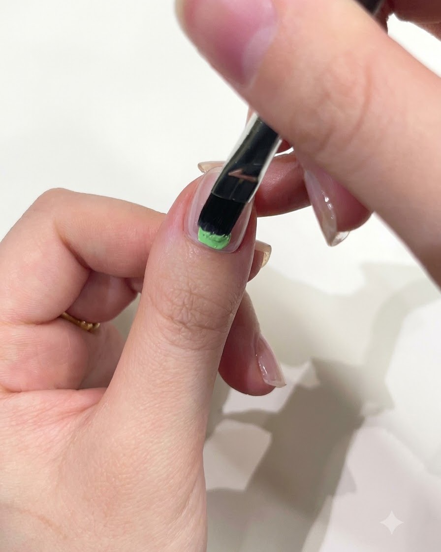 Un primo piano di una mano che utilizza un pennello piatto per applicare il gel solido verde menta alla base dellunghia del pollice.