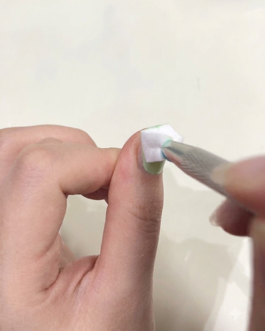 Primo piano di una mano che tiene una pinzetta con una piccola spugnetta bianca per picchiettare il gel verde su ununghia.