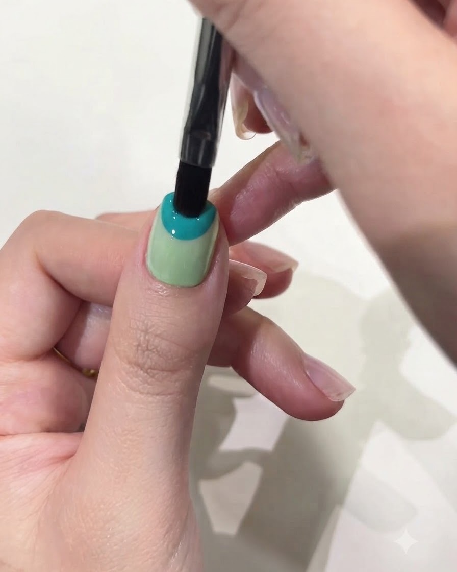 Primo piano di un pennello piatto che applica del gel solido blu pastello sulla metà superiore di ununghia del pollice.
