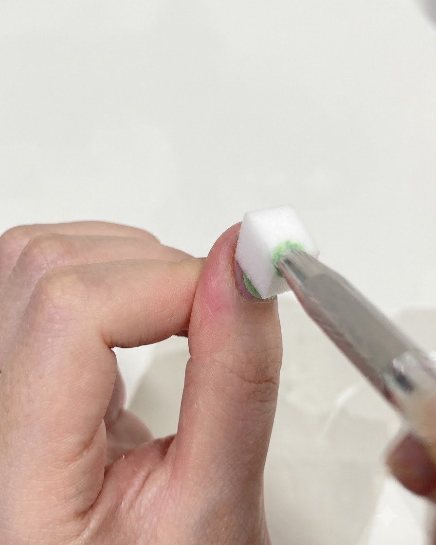 Primo piano di una pinzetta che tiene una piccola spugnetta da trucco bianca, picchiettando delicatamente la superficie di un pollice per unire insieme i gel verde e blu.
