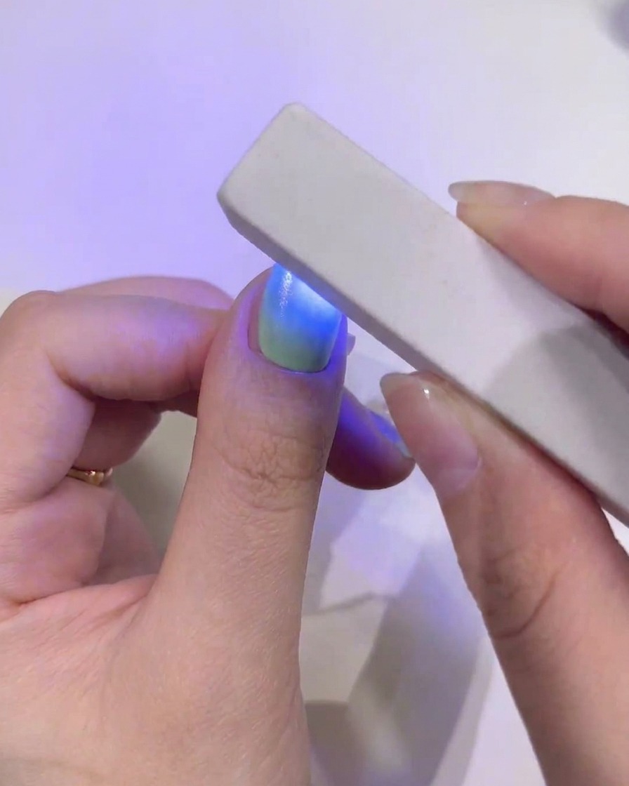 Primo piano di ununghia del pollice con un perfetto gradiente verde e blu che viene polimerizzata sotto una mini lampada UV/LED blu rettangolare e portatile.