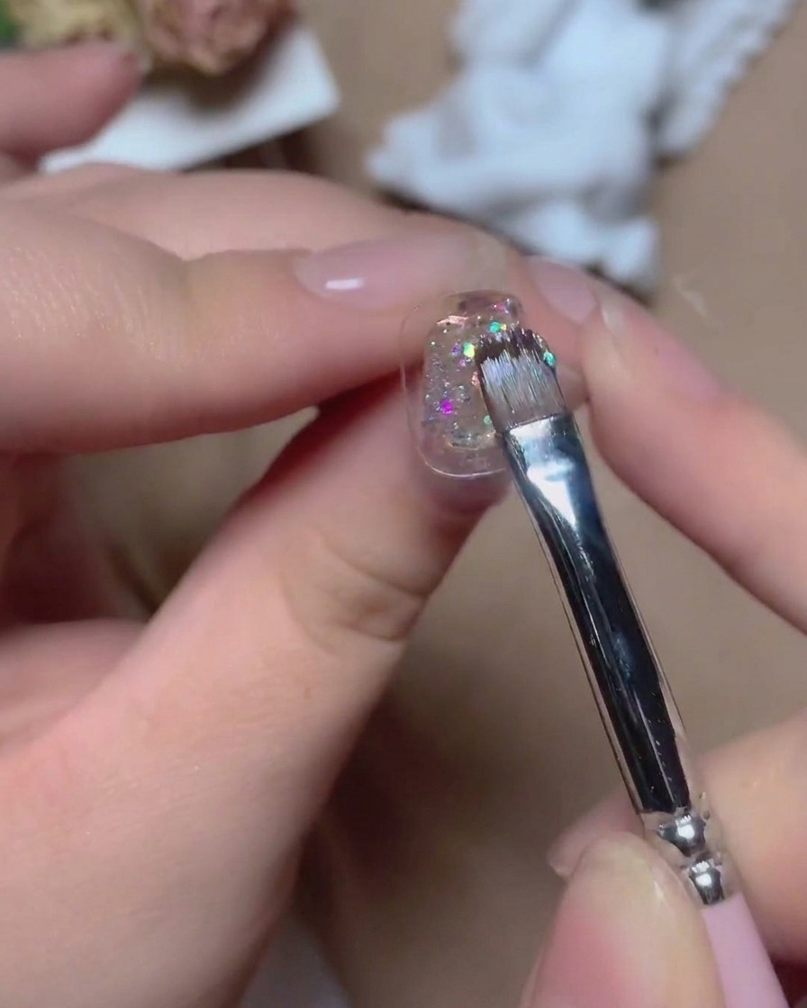 Un pennello sottile applica un gel glitter multicolore brillante su una superficie ungueale con riflessi vibranti.