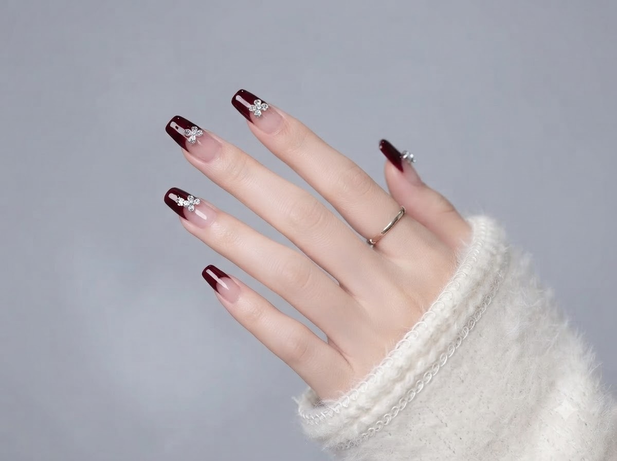 Una mano che mostra la disposizione completa delle unghie in gel bordeaux e nude con dettagli in cristallo.