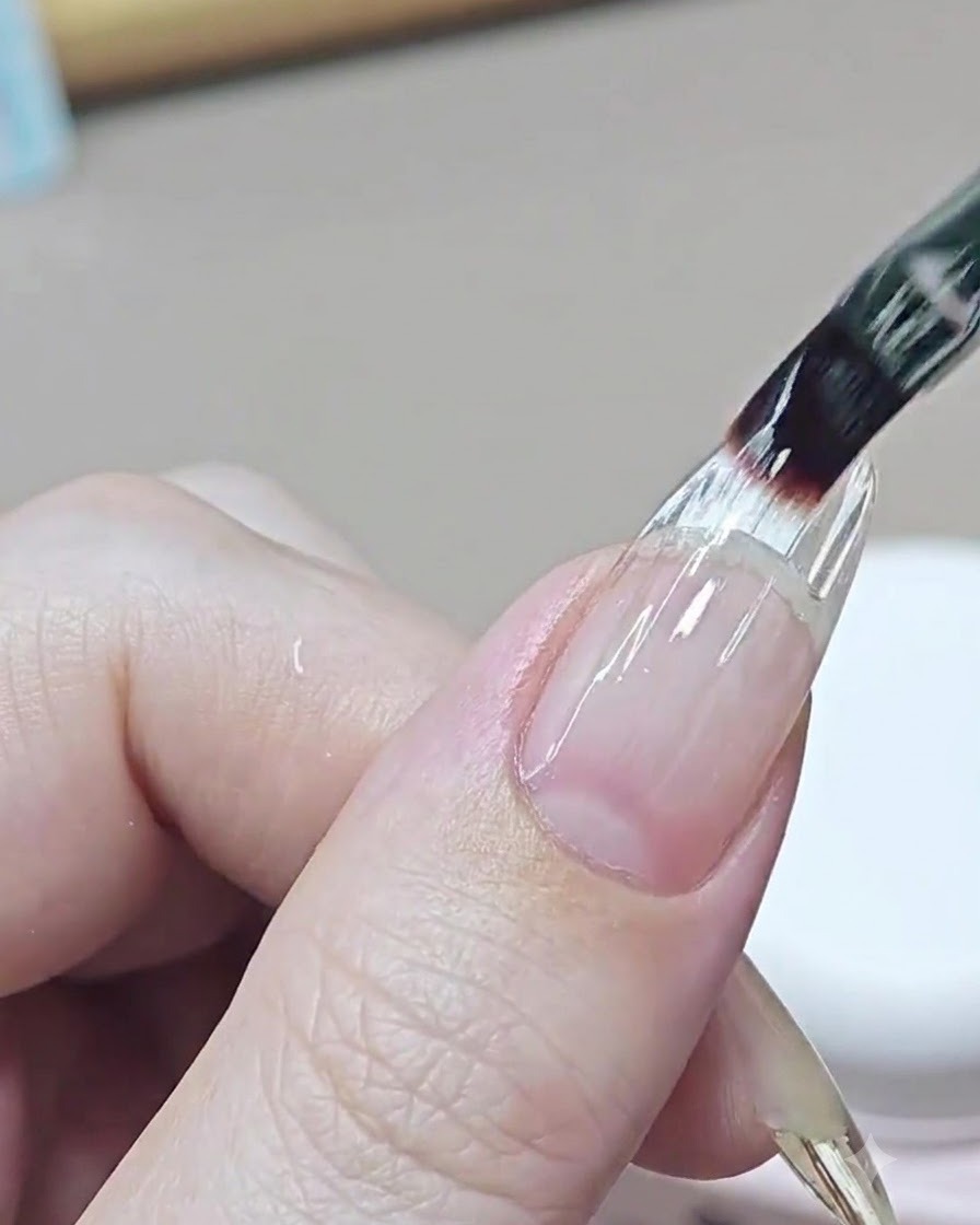 Un pennello fine per unghie applica uno strato trasparente e livellante di gel costruttore sullarea di transizione di ununghia allungata.