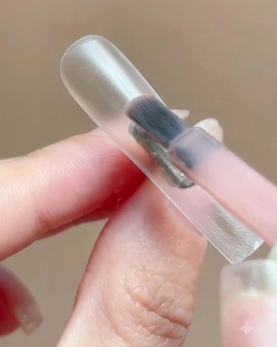 Un pennello che applica un sottile strato di gel polish rosa traslucido su un supporto per unghie trasparente.