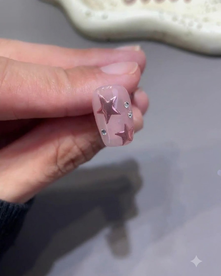 Un minuscolo pennello da dettaglio che dipinge meticolosamente il top coat attorno alla base di piccoli strass su ununghia rosa finita con stelle.