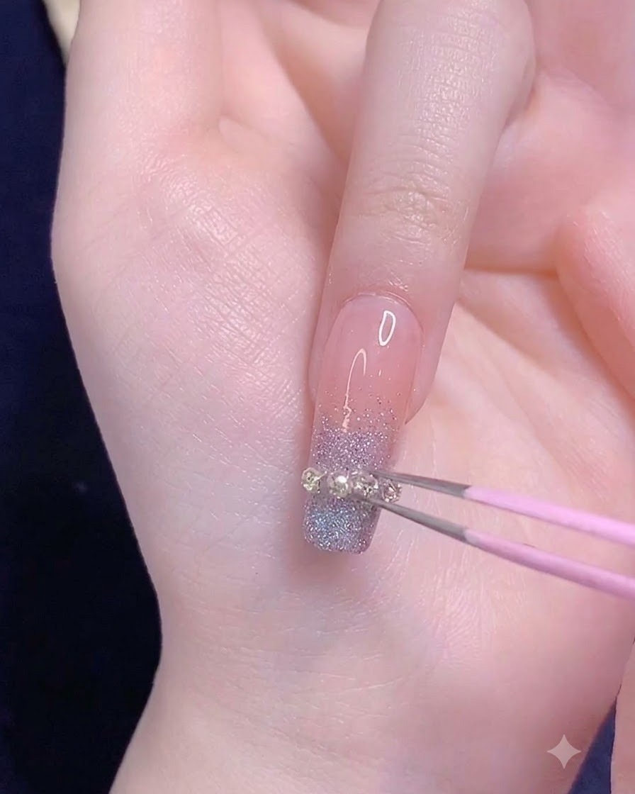 Una pinzetta rosa che posiziona una piccola striscia di catenella metallica in modo orizzontale sulla punta sfumata di ununghia anulare.