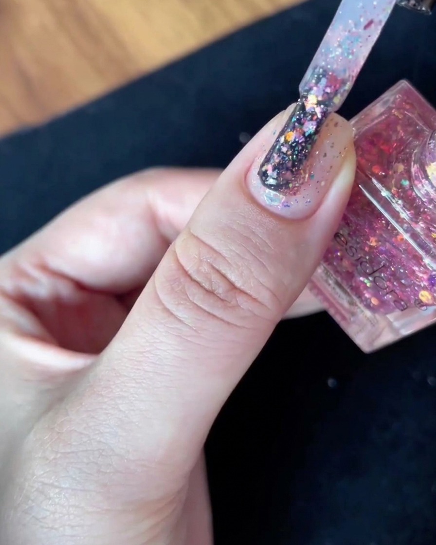 Un pennello da smalto pieno di scaglie di glitter tenuto fermo appena sopra la superficie di ununghia con base nudo.