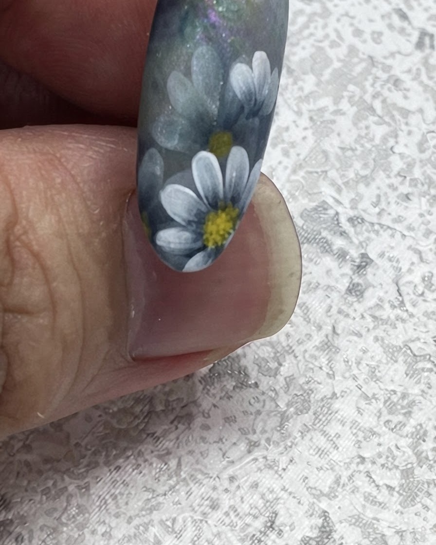 Primo piano di un pennello per unghie che tira verso lesterno per creare una punta fogliare affilata accanto a margherite dipinte in stile nordico.